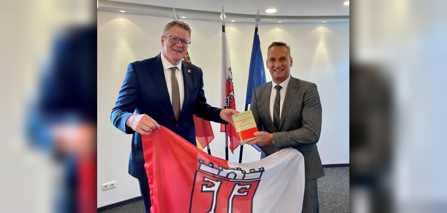 Das Foto zeigt Jörn Patzak, den Leiter der JVa Wittlich und Bürgermeister Rodenkirch bei der Übergabe der Fahne der Stadt Wittlich.